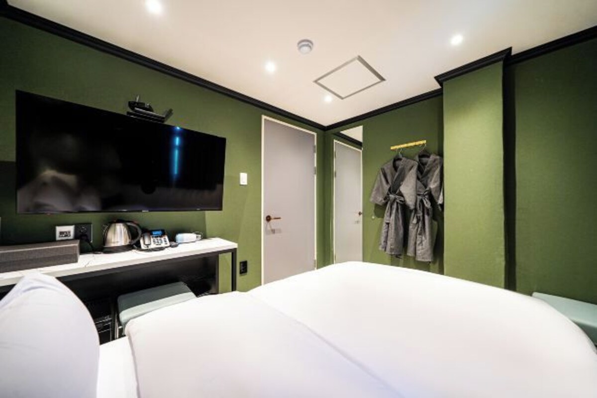 Una habitación de hotel con cama, televisor montado en la pared, escritorio con hervidor y armario con ropa colgada.