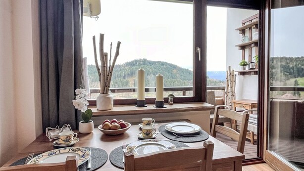 A dining area with a table set for four, a window with a view of a forested mountain, and a shelf with decorative items. - Apartment Wolf by Interhome (Feldkirchen in Kärnten)