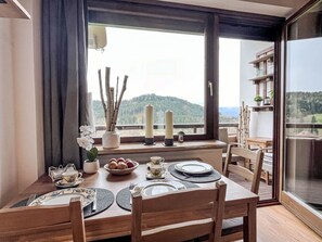 A dining area with a table set for four, a window with a view of a forested mountain, and a shelf with decorative items. - Apartment Wolf by Interhome (Feldkirchen in Kärnten)