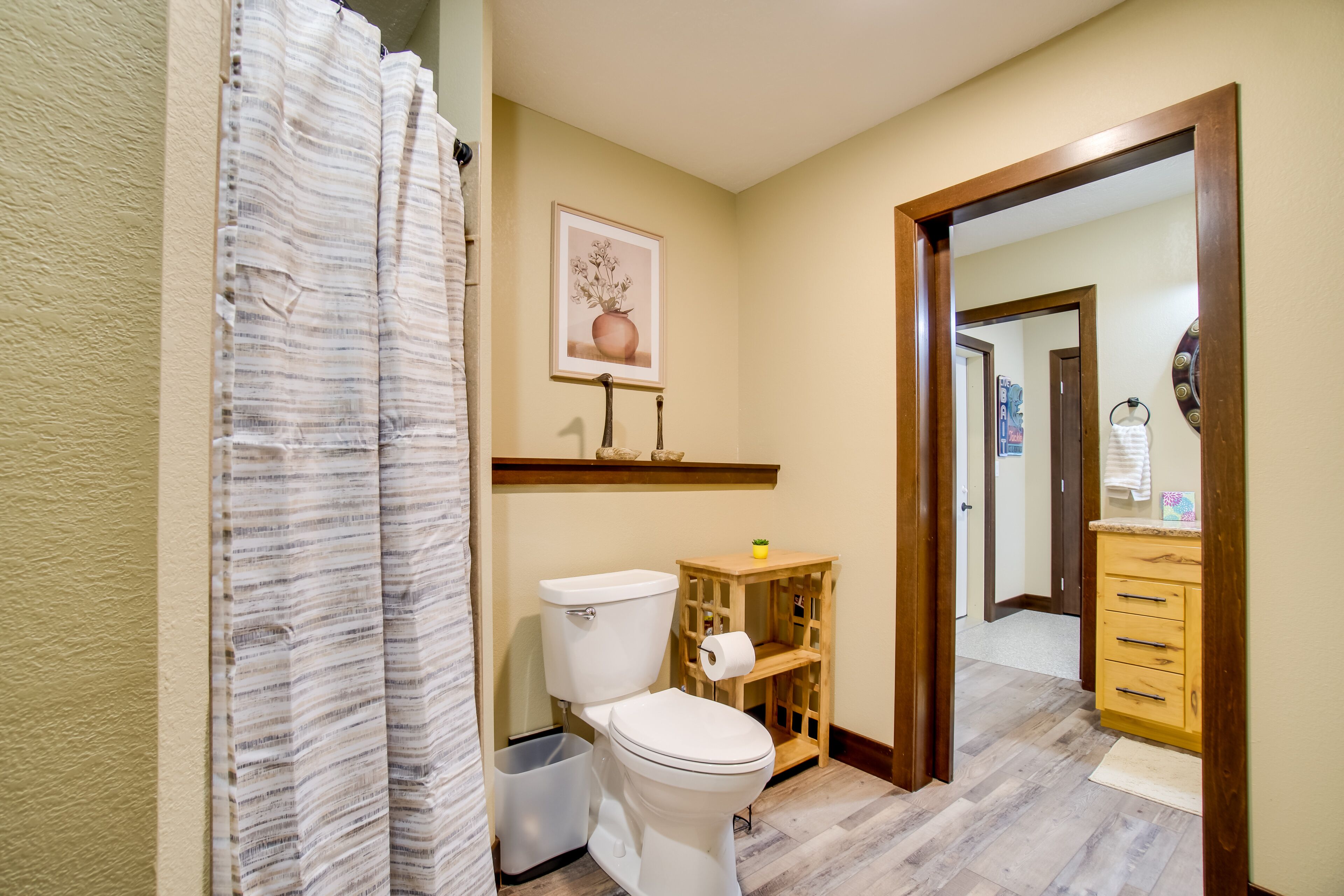 A bathroom with a toilet, a wooden shelf, a trash bin, and a shower curtain.
