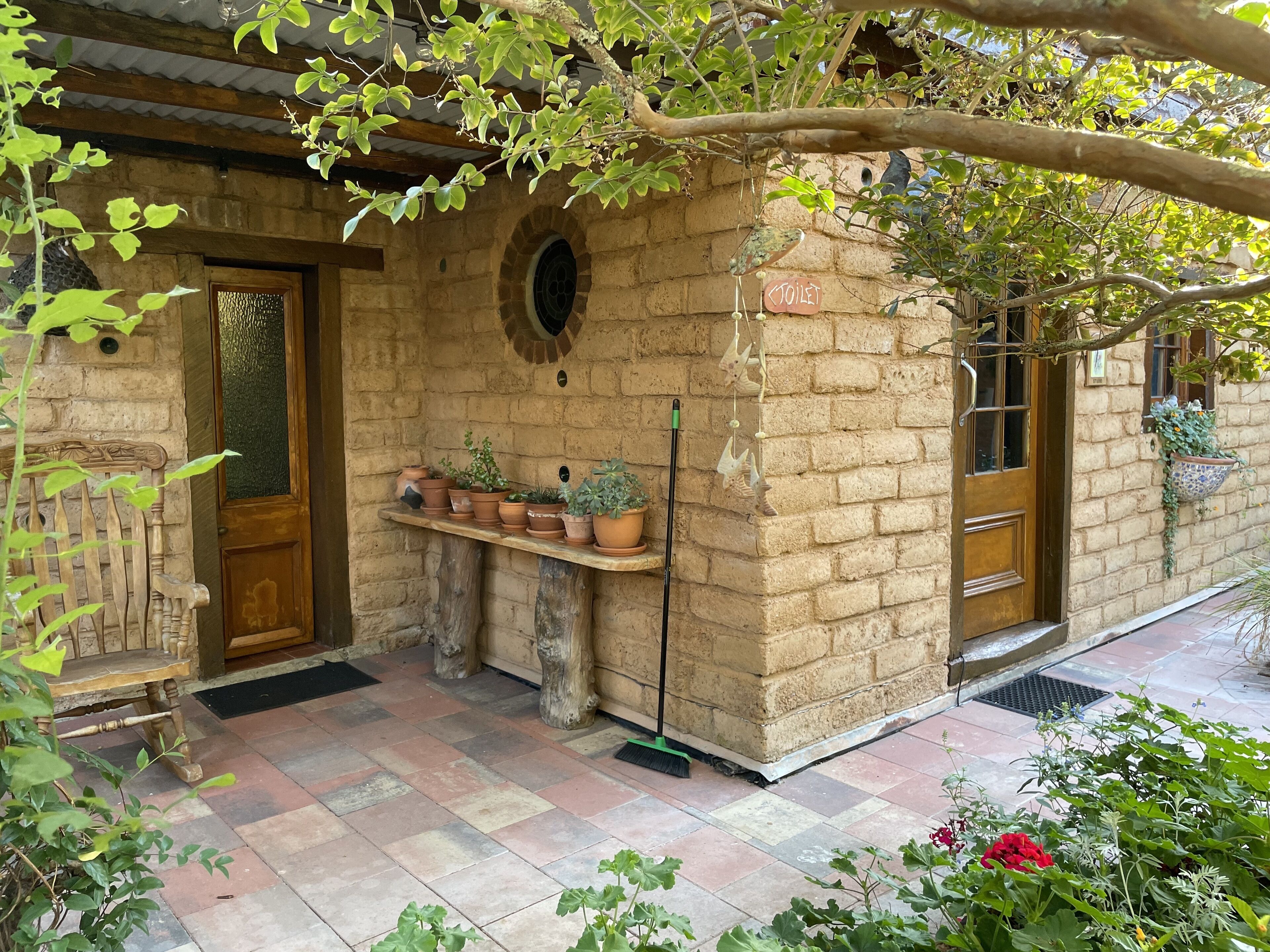 A rustic stone building with a wooden door, a bench, and a shelf with potted plants.