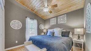 Une chambre avec un plafond en bois, un lit recouvert d’une courtepointe bleue, une chaise blanche et des œuvres d’art décoratives accrochées au mur.
