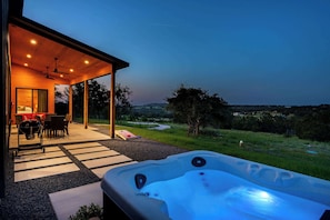 A hot tub with a view of a grassy landscape, a covered patio area with outdoor furniture, and a building with a warm interior light. - Outbound Ranch, Hot tub, Sauna, Fireplace, Views! (fredericksburg)