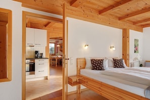 Quarto com estrutura de madeira, cama, mesas de cabeceira e vista para a cozinha.