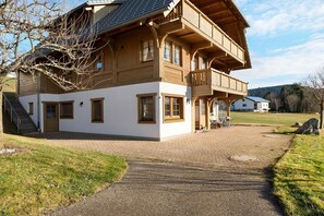 A two-story wooden house with a balcony, a paved driveway, and a grassy yard.