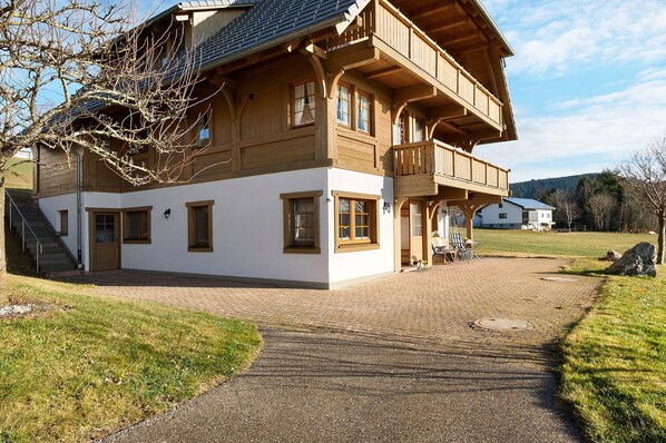 A two-story wooden house with a balcony, a paved driveway, and a grassy yard.