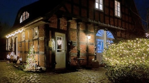 Un bâtiment traditionnel à colombages, avec un chemin pavé menant à l’entrée, décoré de guirlandes lumineuses et d’un petit sapin de Noël.