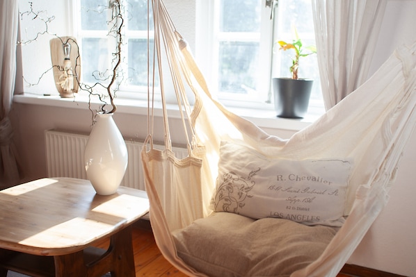 Une pièce avec un fauteuil suspendu, une table, un vase et une fenêtre avec des rideaux.