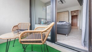 A room with a glass door leading to a balcony with wicker chairs and a small table. Inside, there is a bed and a television mounted on the wall.