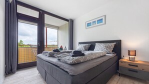 A bedroom with a large bed, a nightstand, a lamp, and a view of the outdoors.