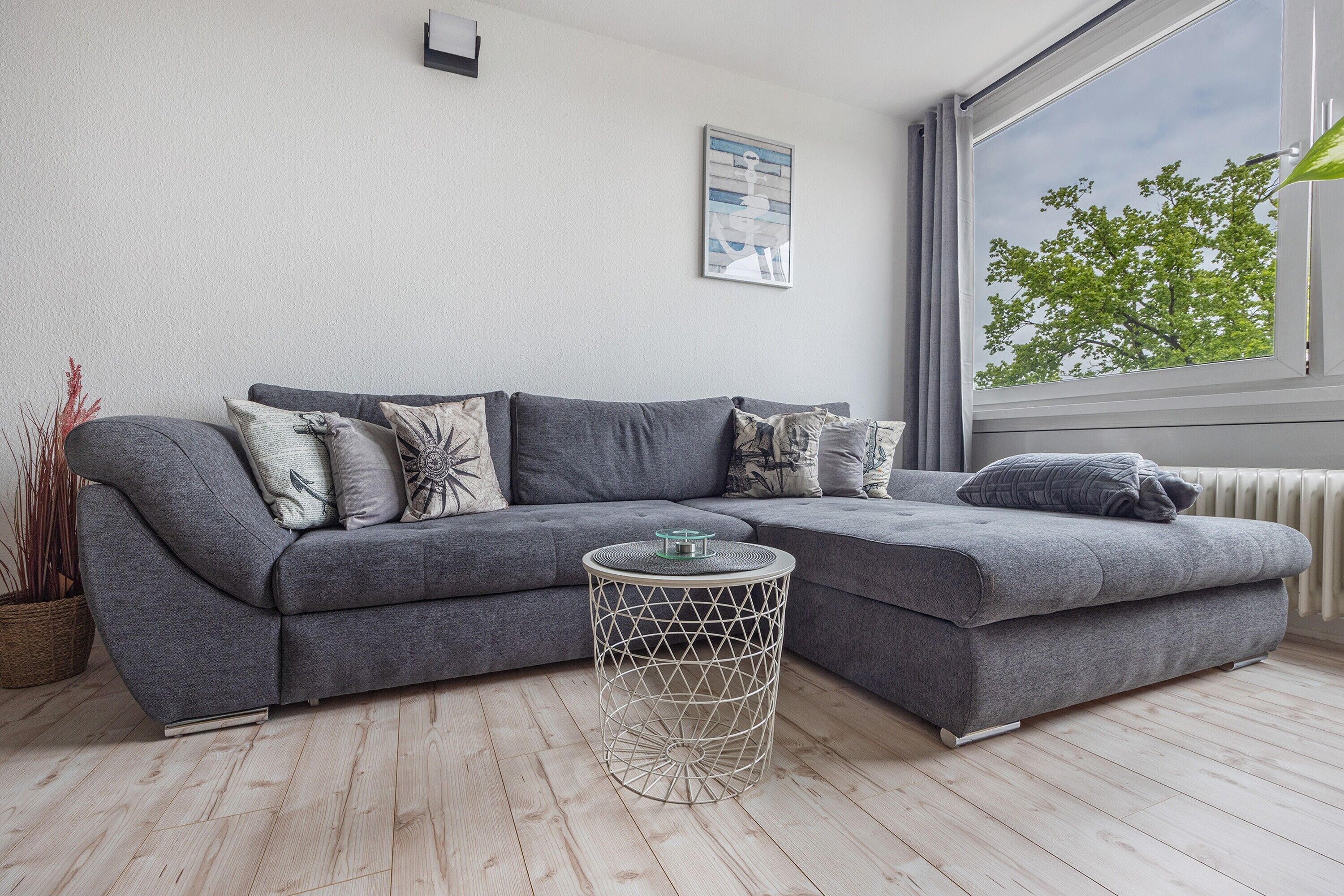 Un salon moderne avec un canapé d’angle gris, une petite table ronde et une grande fenêtre donnant sur la forêt.