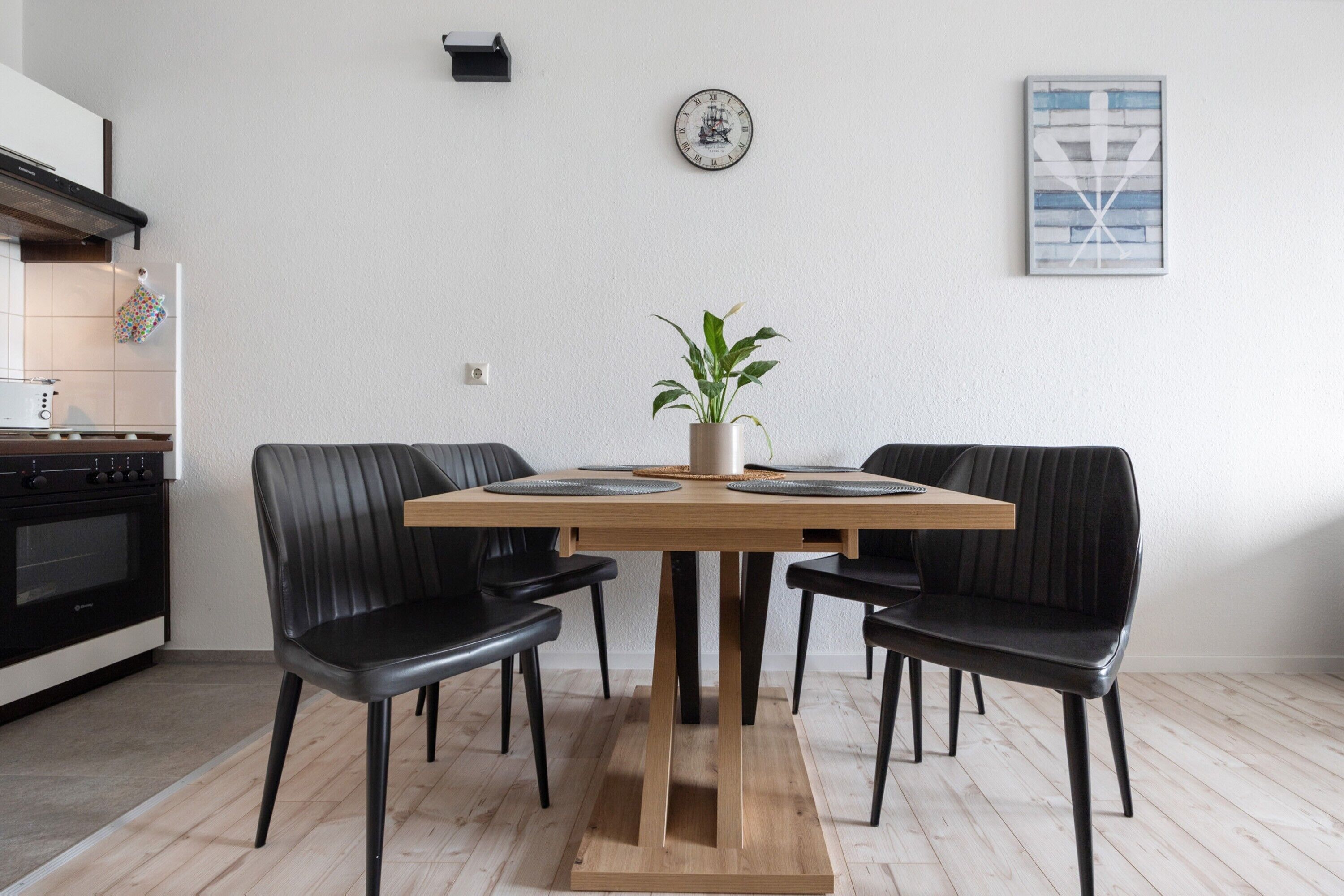 A dining area with a wooden table, black chairs, a potted plant, a clock, and a framed picture on the wall.