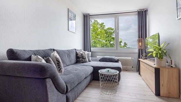 A modern living room with a grey sofa, a round coffee table, a wooden sideboard, and a large window with a view of trees.