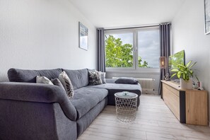 A modern living room with a grey sofa, a round coffee table, a wooden sideboard, and a large window with a view of trees.