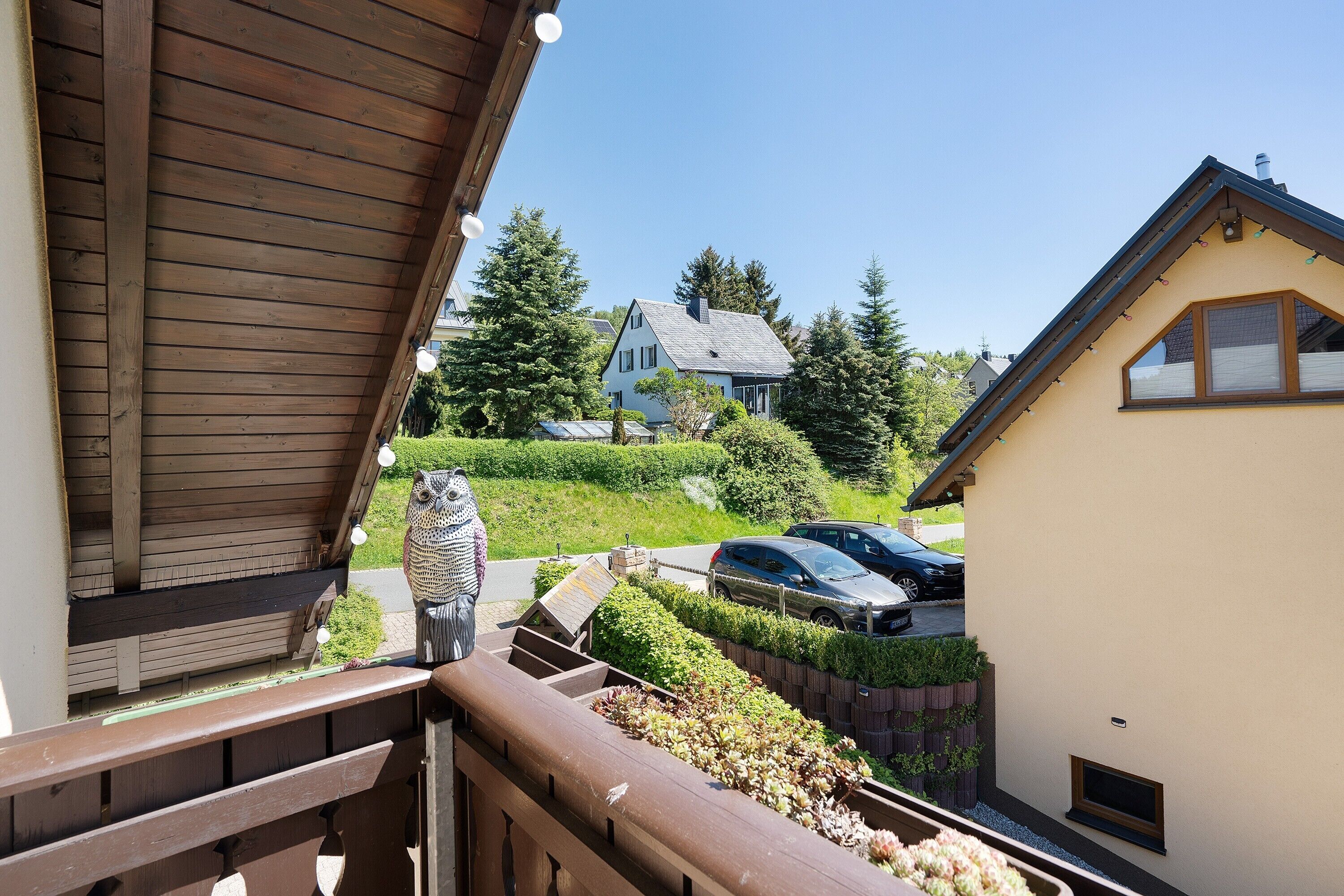 A balcony with a wooden railing overlooking a residential street, a parked car, and a house with a sloped roof.