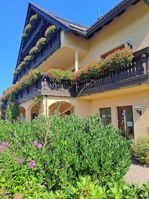 Un immeuble à plusieurs étages, doté de balcons ornés de fleurs rouges, d’une entrée accueillante et d’un jardin bien entretenu.
