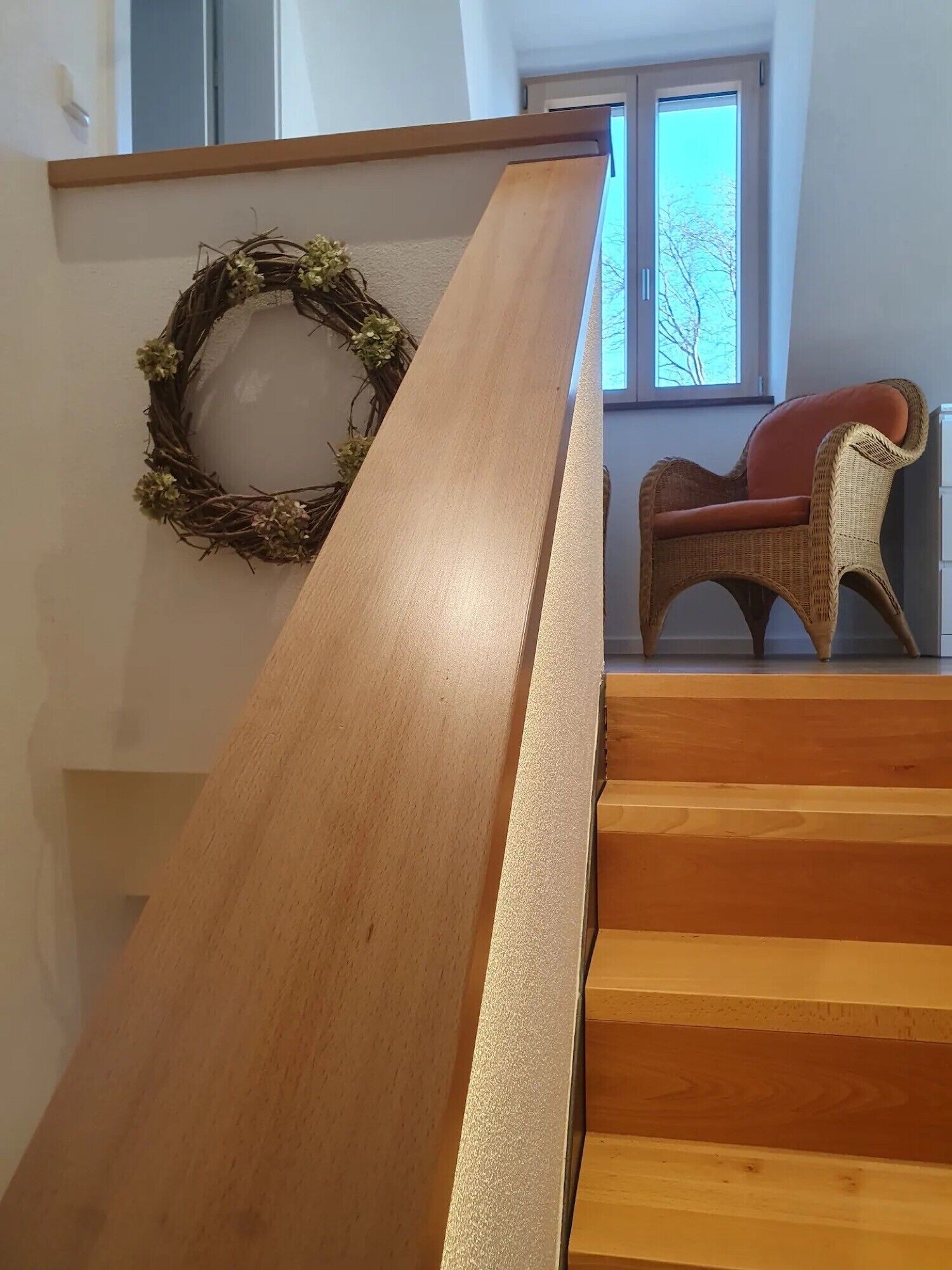 Eine Holztreppe mit Geländer, ein Korbstuhl und ein Kranz an der Wand.