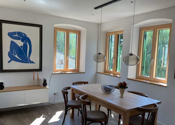 A dining area with a wooden table, chairs, pendant lights, and a framed artwork on the wall.