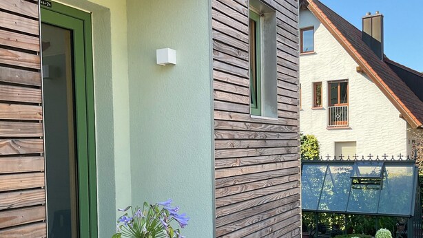 A modern house with a green door, wooden siding, and a small garden with flowers.