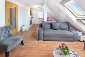 Ein modernes Apartment mit grauem Sofa, einem gesteppten Sessel, Holzboden, einer Dachfensterkuppel und einer Küche im Hintergrund.