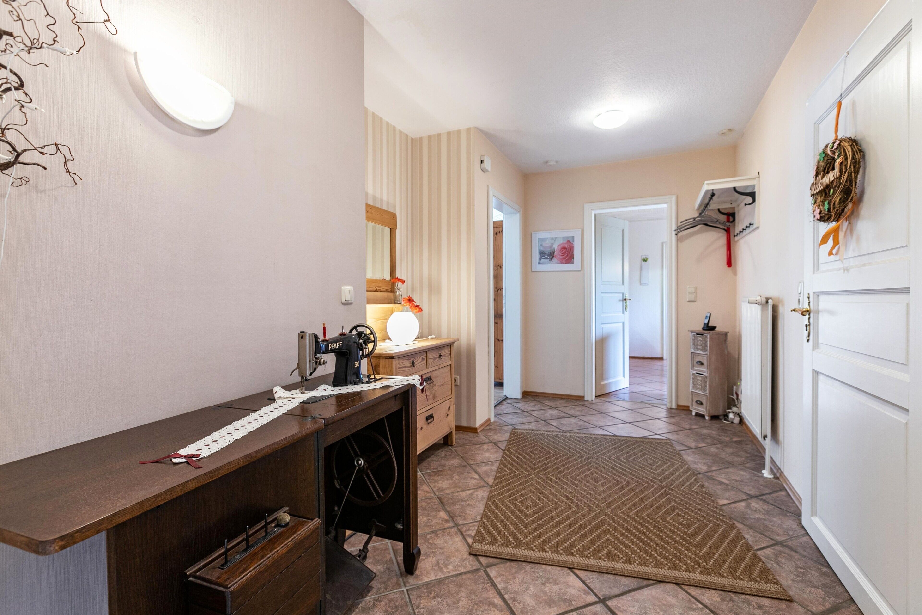 Un couloir avec une machine à coudre, une lampe, un tapis et une porte ornée d’une guirlande.