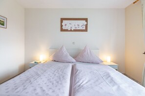 Ein ordentlich bezogenes Bett mit grau-weißer Spitze, zwei Nachttischlampen und einem gerahmten Blumenbild an der Wand.