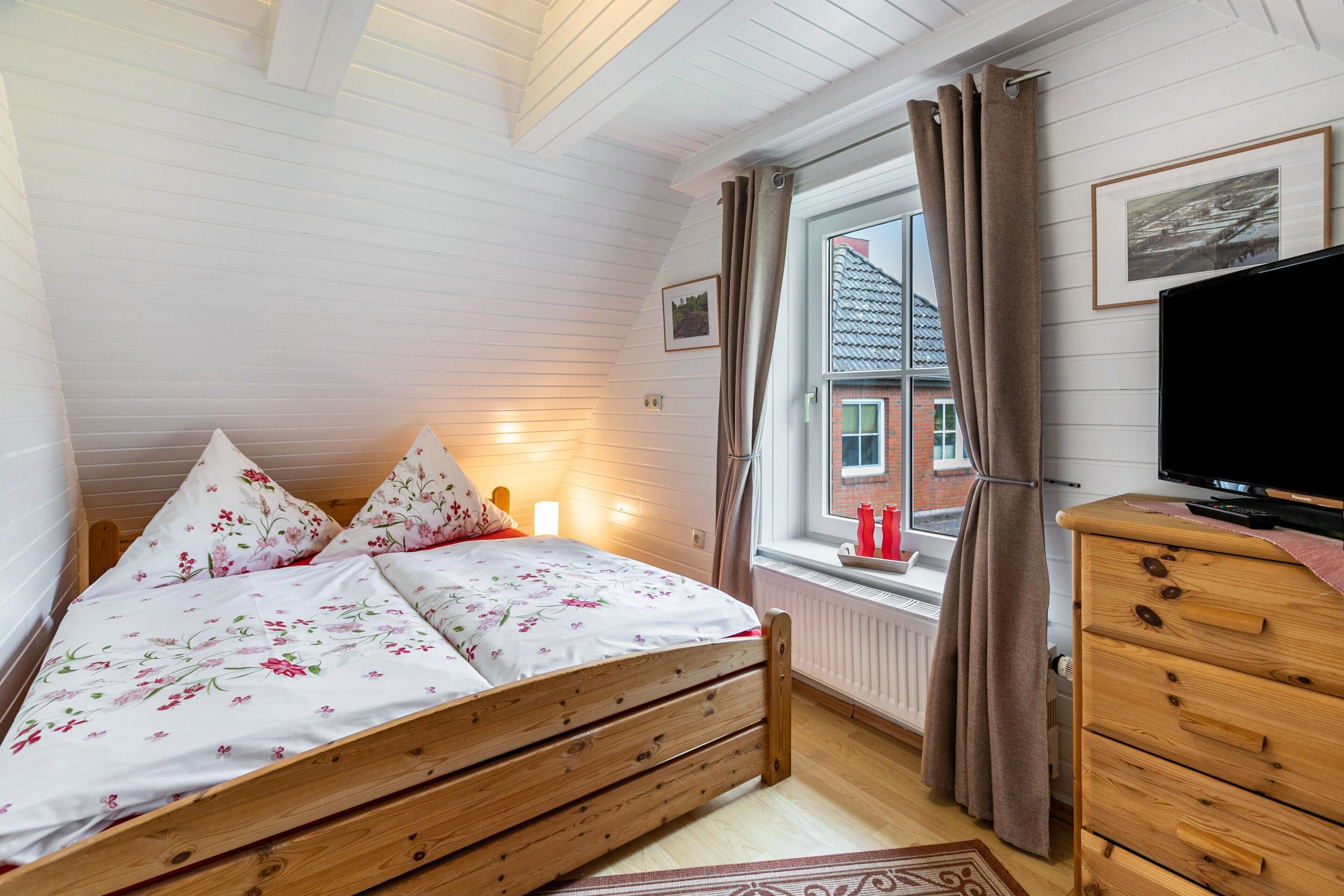 Quarto com cama de madeira, cômoda, televisão, janela com cortinas e radiador.