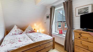 Quarto com cama de madeira, cômoda, televisão, janela com cortinas e radiador.