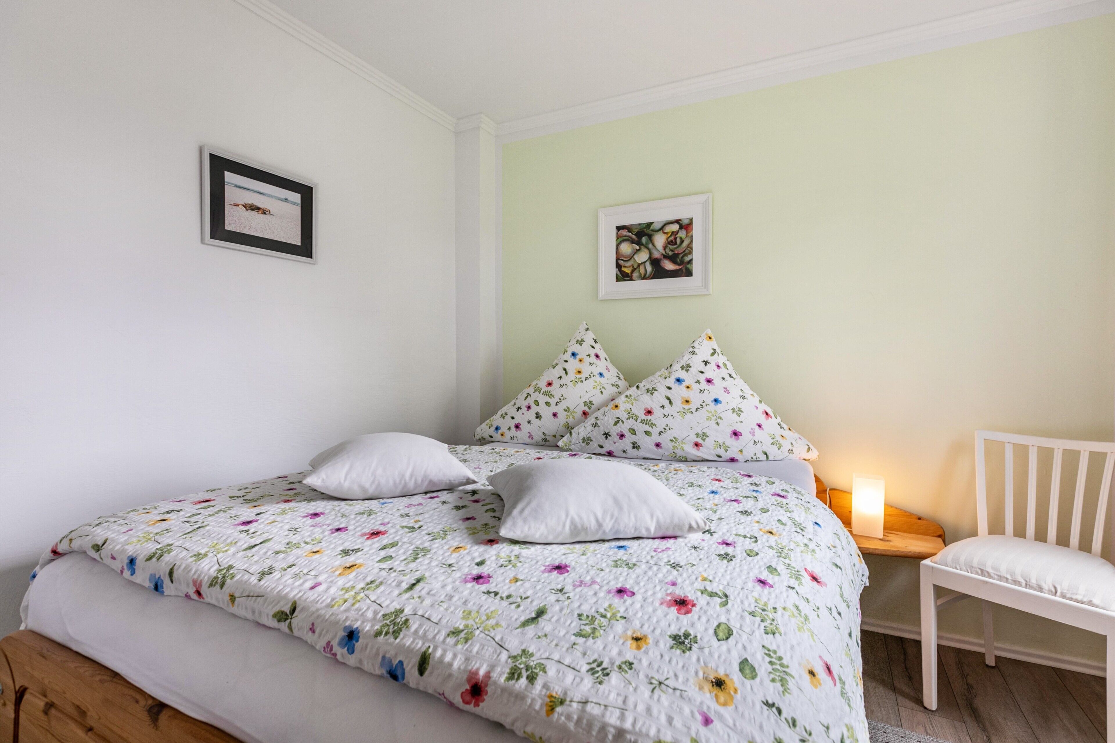 A bed with a floral quilt, white pillows, a white chair, and a bedside table with a lamp.