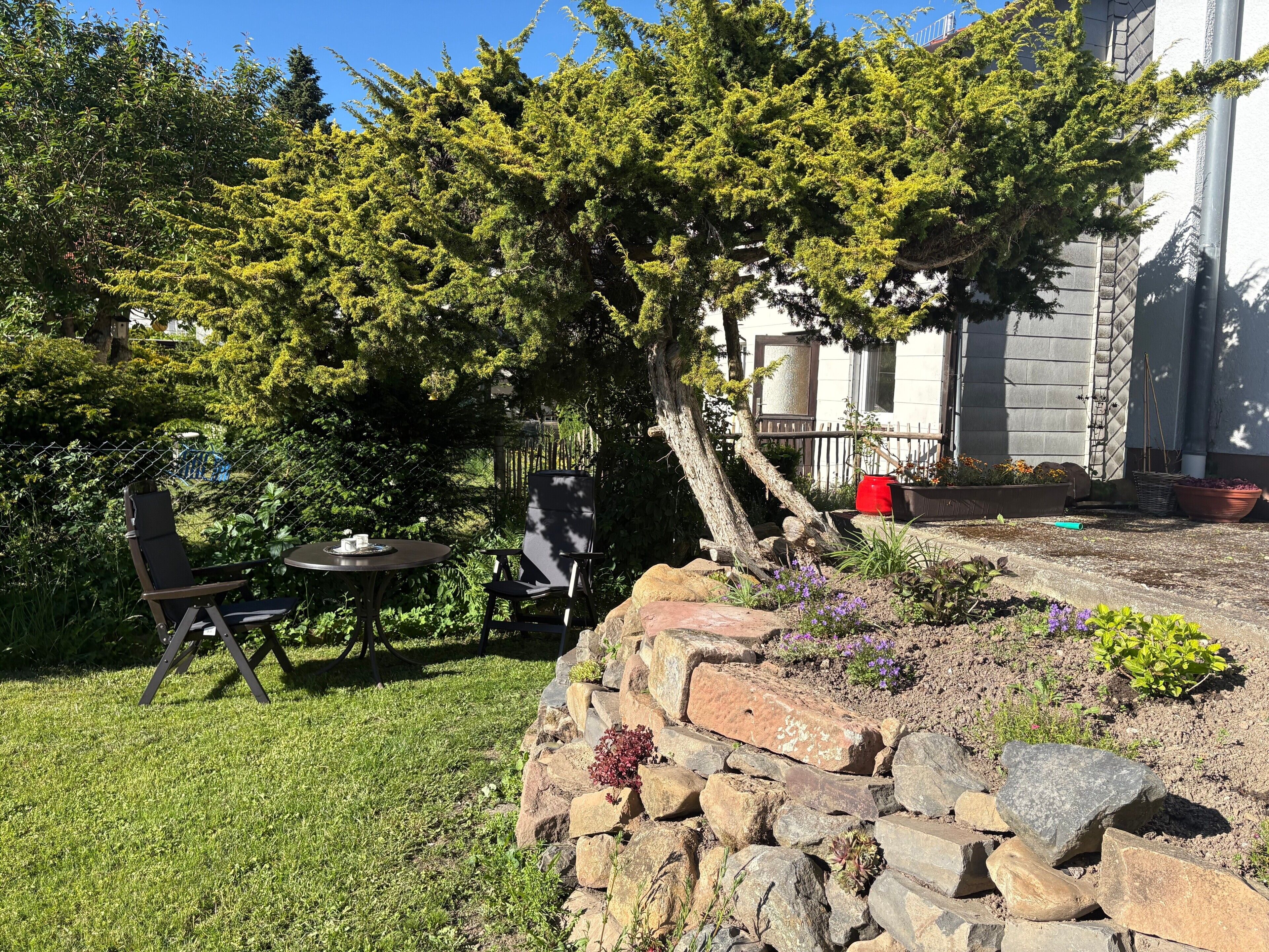 A stone retaining wall, a small table with two chairs, and a tree in a garden area.
