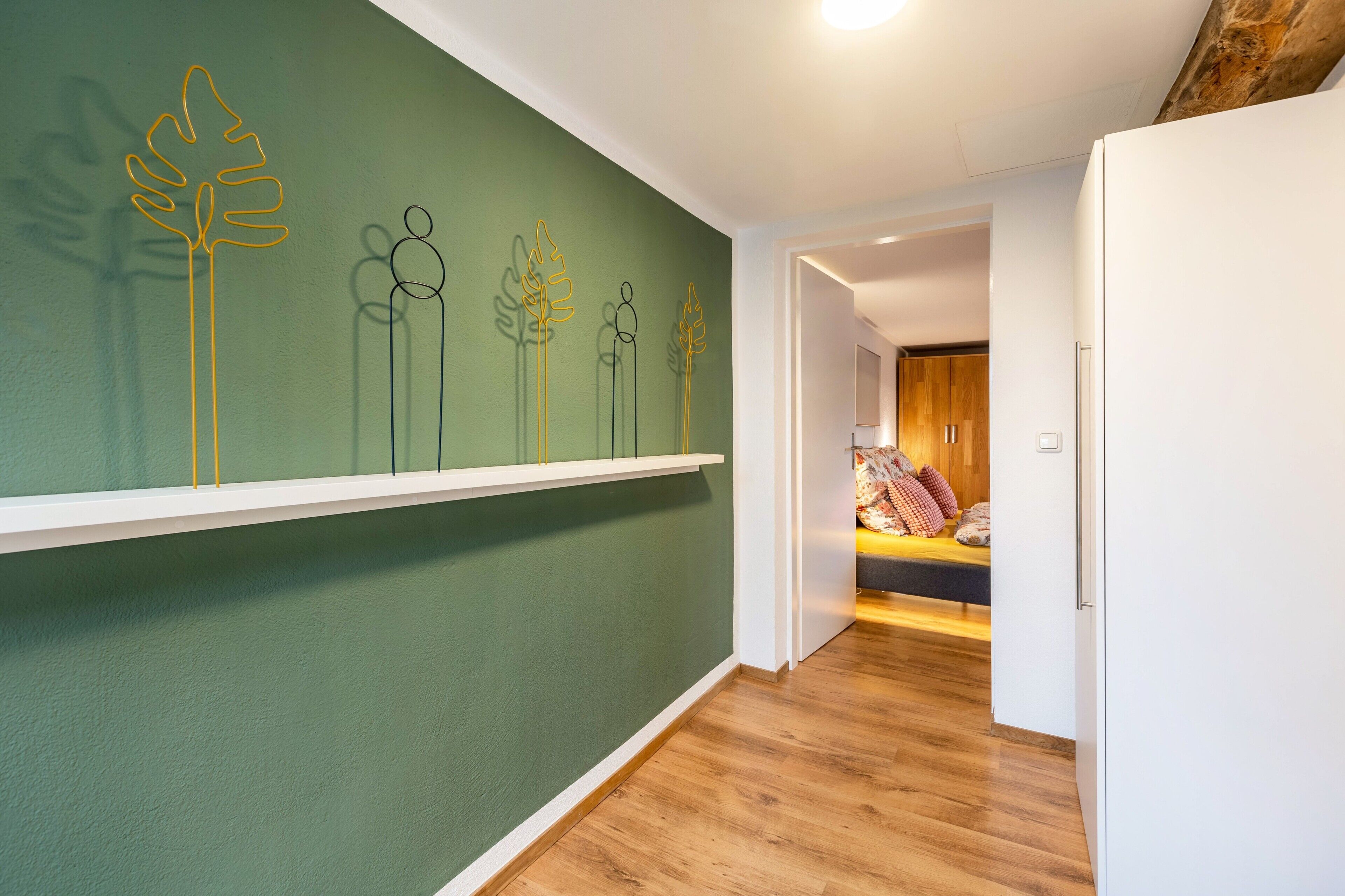 Un corridoio con parete verde, pavimento in legno e una porta bianca che conduce a una camera da letto con letto e cuscini.