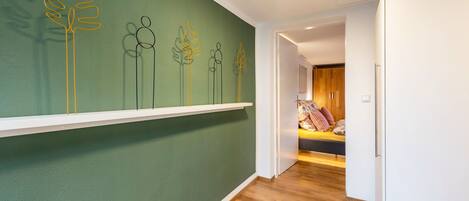 A hallway with a green wall, wooden flooring, and a white door leading to a bedroom with a bed and pillows.