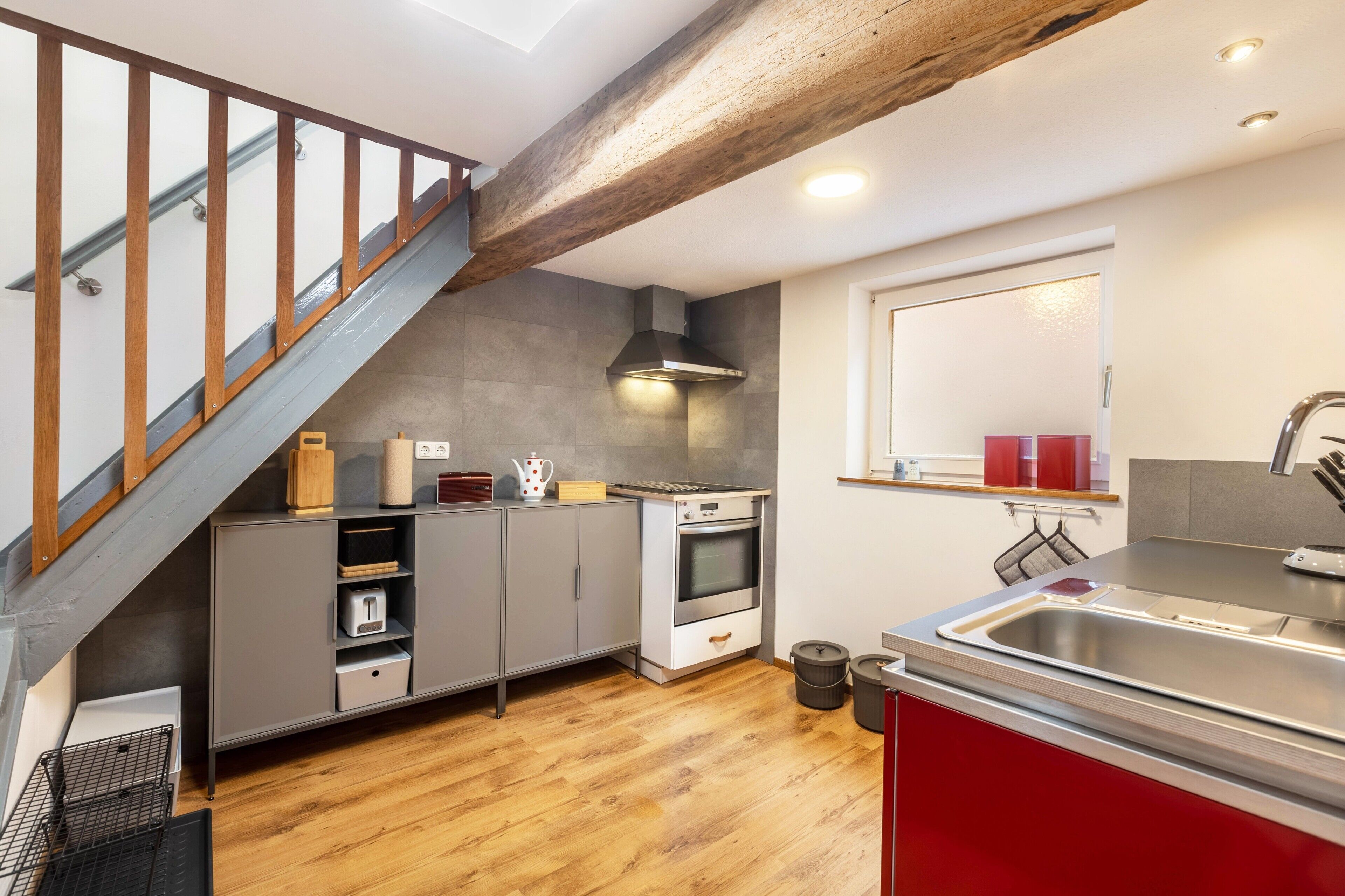 Una cocina con escalera de madera, armarios grises, un lavamanos de acero inoxidable y una ventana con cortinas rojas.