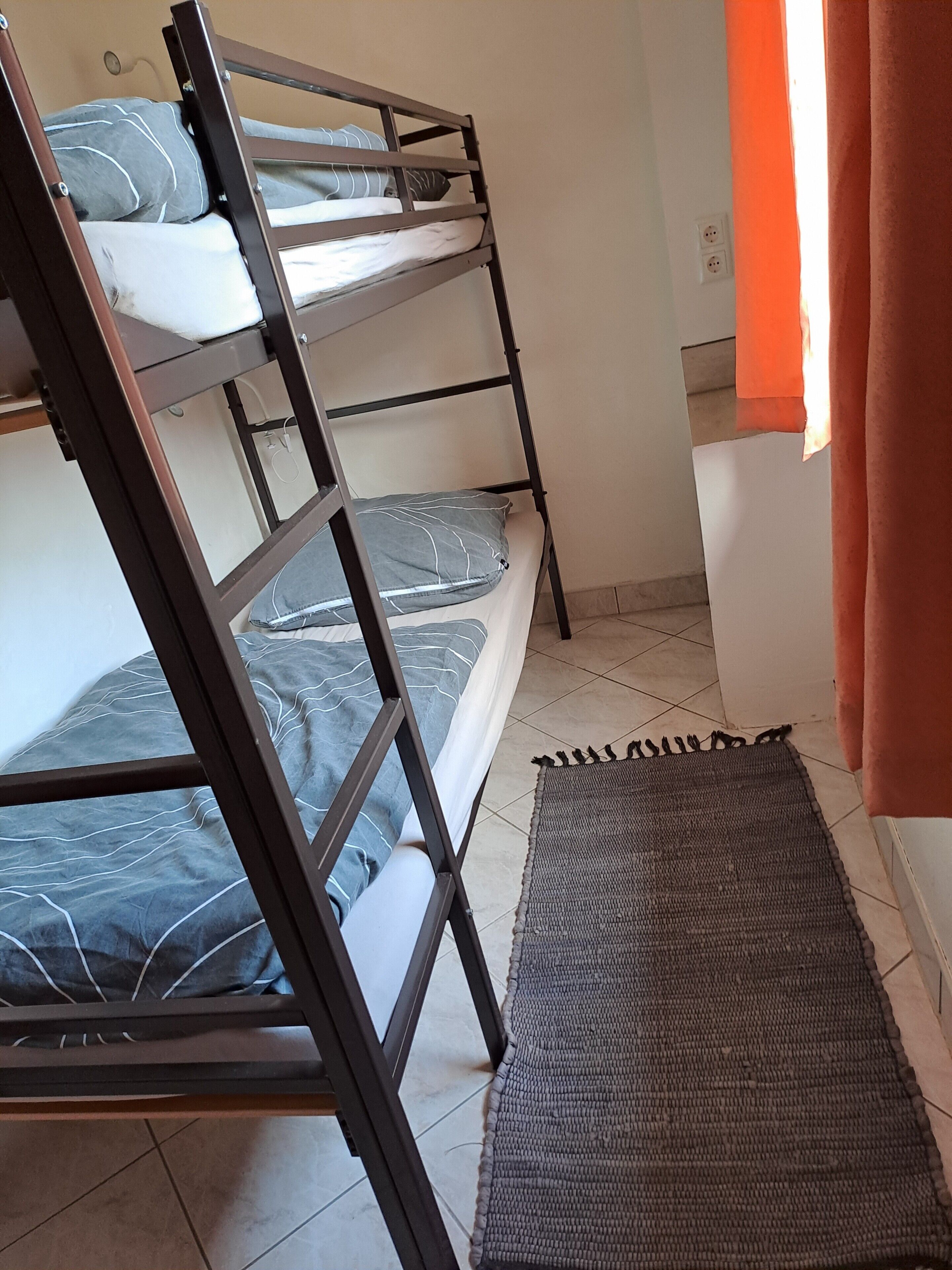 Ein Etagenbett mit grauer Bettwäsche, einer gewebten Matte und einem Badezimmer mit Waschbecken in Weiß und orangefarbenen Vorhängen.
