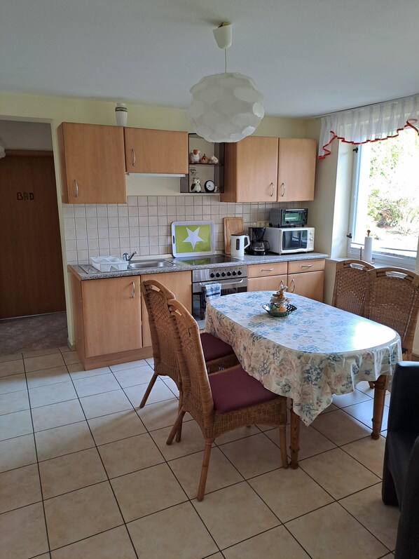 A kitchen with wooden cabinets, a dining table with a floral tablecloth, wicker chairs, and a view of the outdoors.
