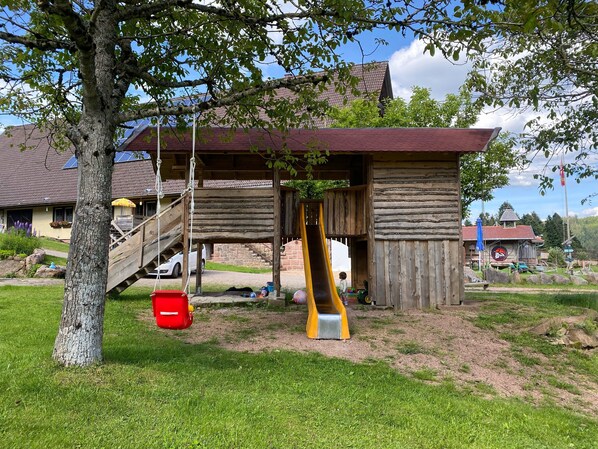 A wooden playhouse with a slide, a hanging swing, and a red bucket.