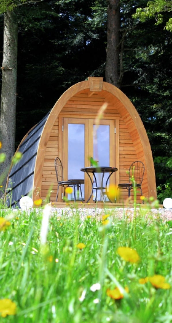 Una cabana de fusta amb dues cadires i una taula, envoltada per herba i arbres.