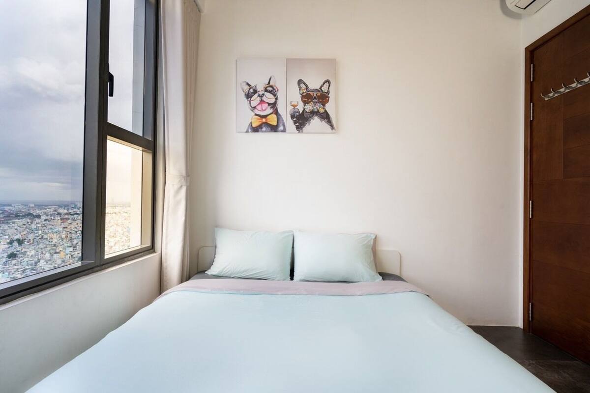 A bedroom with a bed, two pillows, a window with a city view, and two wall art pieces.