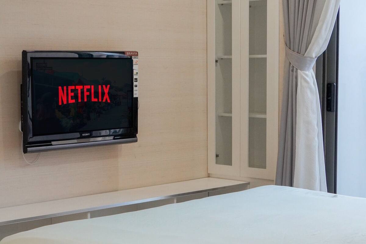 A hotel room with a bed, a TV mounted on the wall displaying 'NETFLIX', a bedside table with a remote control, and a window with curtains.