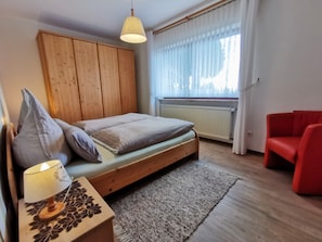 A bedroom with a wooden bed, a red chair, a bedside table with a lamp, and a window with curtains. - Apartment 'Zur Schönen Aussicht' with Mountain View, Private Terrace and Wi-Fi (Schotten)
