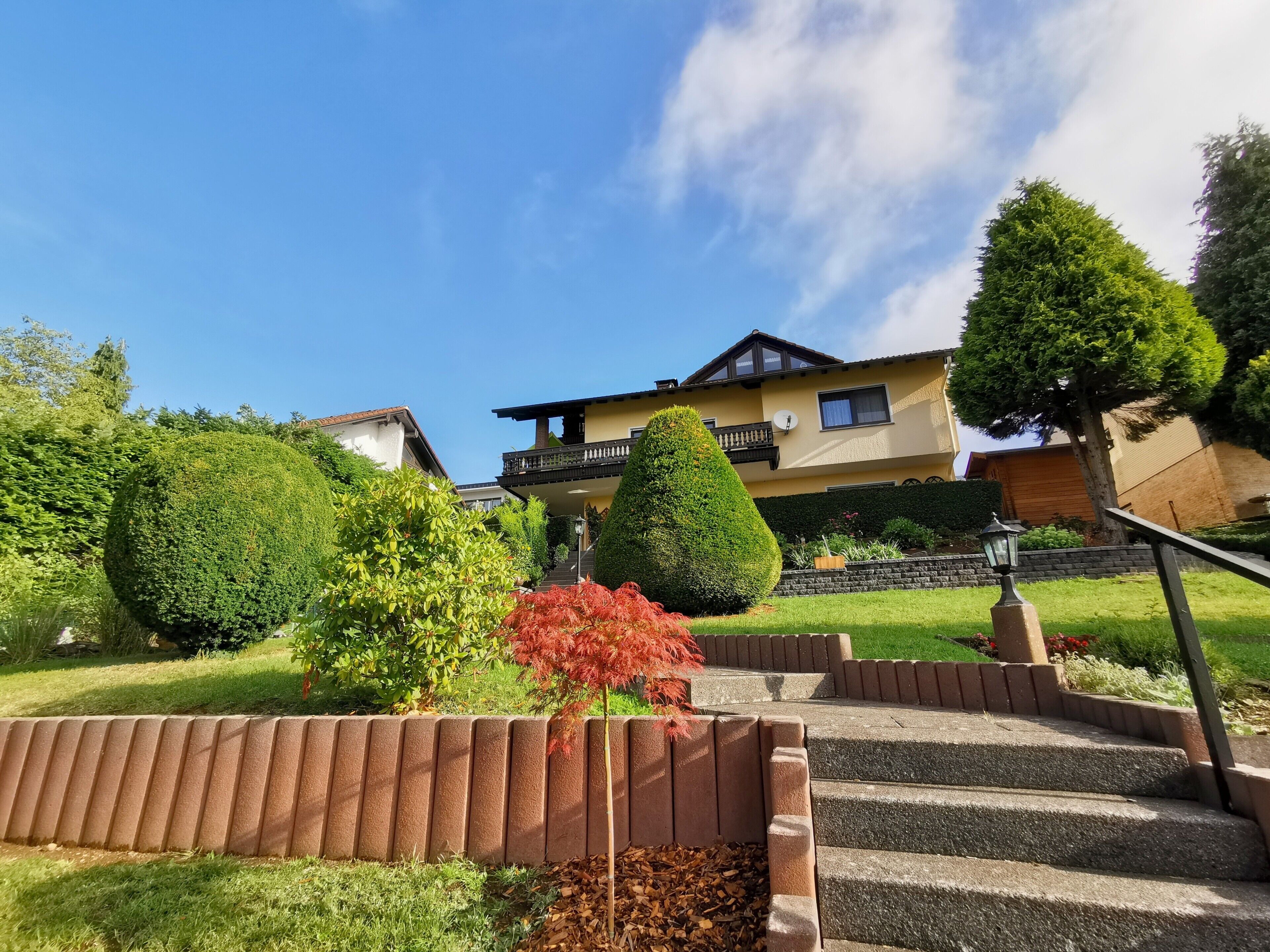 A two-story house with a balcony, surrounded by well-maintained gardens and a clear sky.