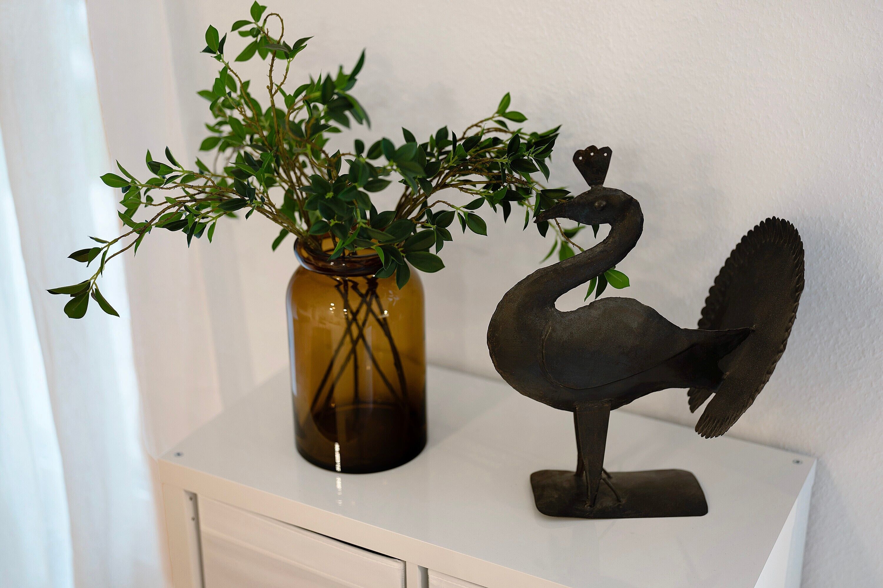 Eine Vase mit grünem Blattwerk und einer Schwane aus Metall auf einem weißen Tisch vor einer weißen Wand.