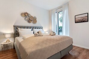 A bedroom with a large bed, a bedside table with a lamp, a decorative wall piece, and a window with curtains.