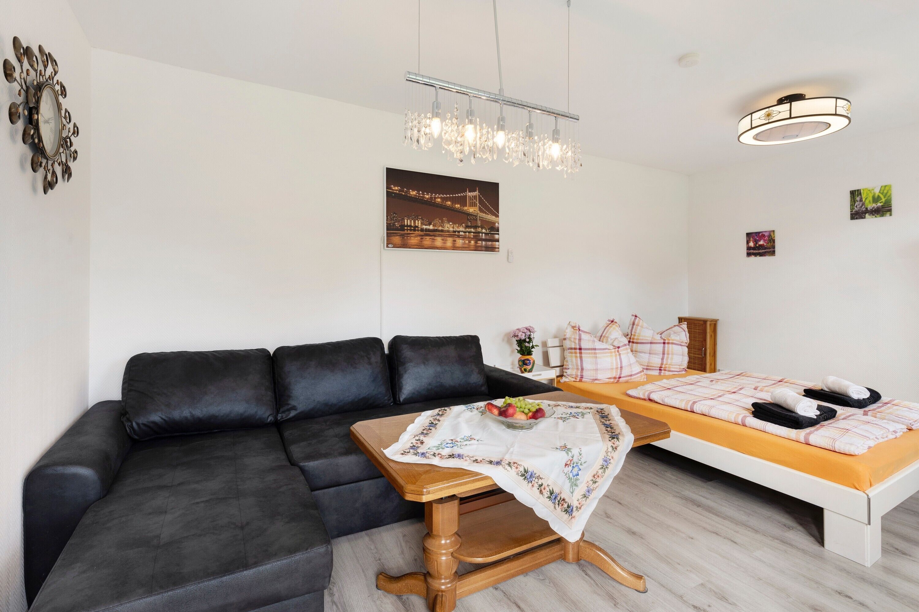 A modern room with a black leather sofa, a wooden table with a fruit bowl, a bed with a checkered blanket, and a chandelier.
