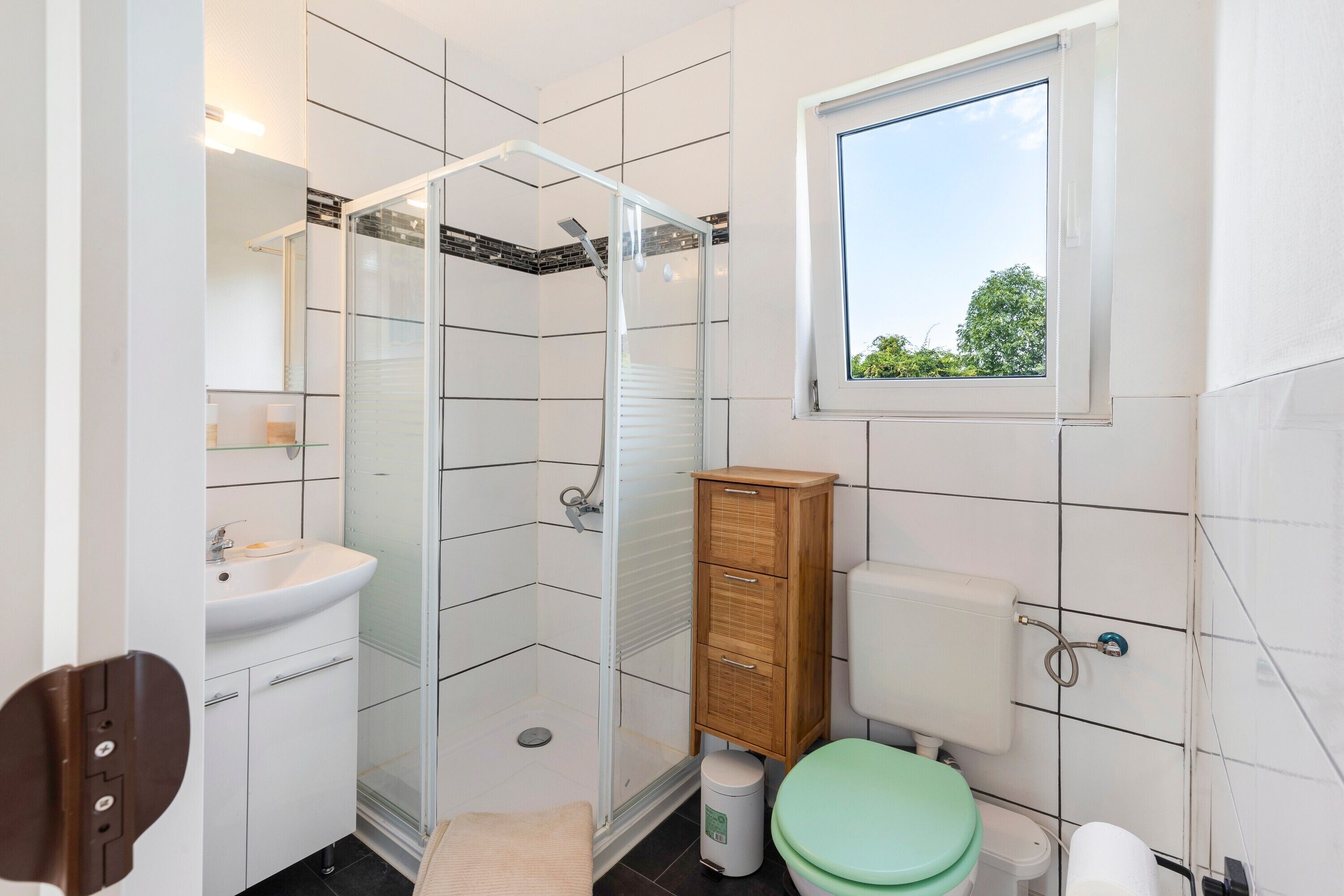 Een badkamer met een douchecabine van glas, een wastafel, toilet, kast en raam.