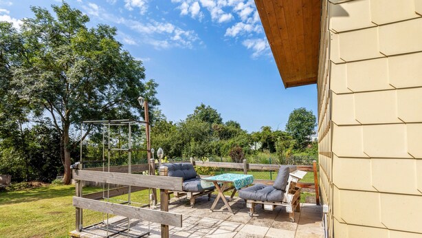 An outdoor patio with a wooden pergola, two grey cushioned chairs, a small table with a blue tablecloth, and a wooden bench.