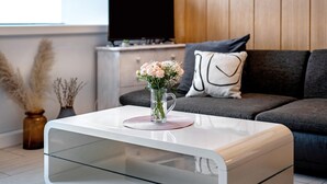 A modern living room with a glass coffee table, a vase with flowers, a flat-screen TV, and a sofa with a decorative pillow. - Apartment 'Ferienwohnung Alpensicht' with Lake View, Shared Garden and Wi-Fi (Oberteuringen)