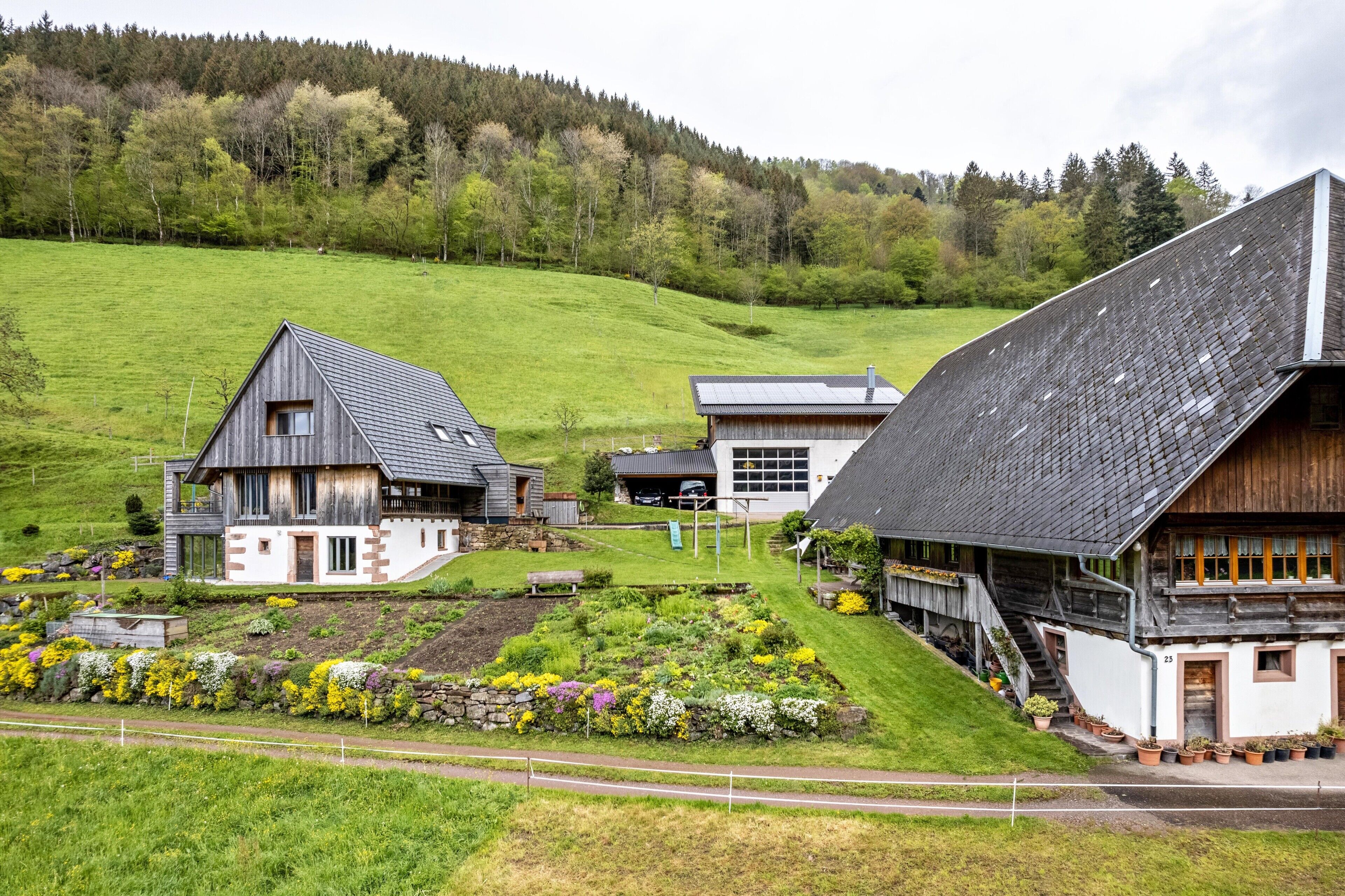 Een landelijke omgeving met een traditioneel houten huis, een modern gebouw en een weelderig groen landschap.