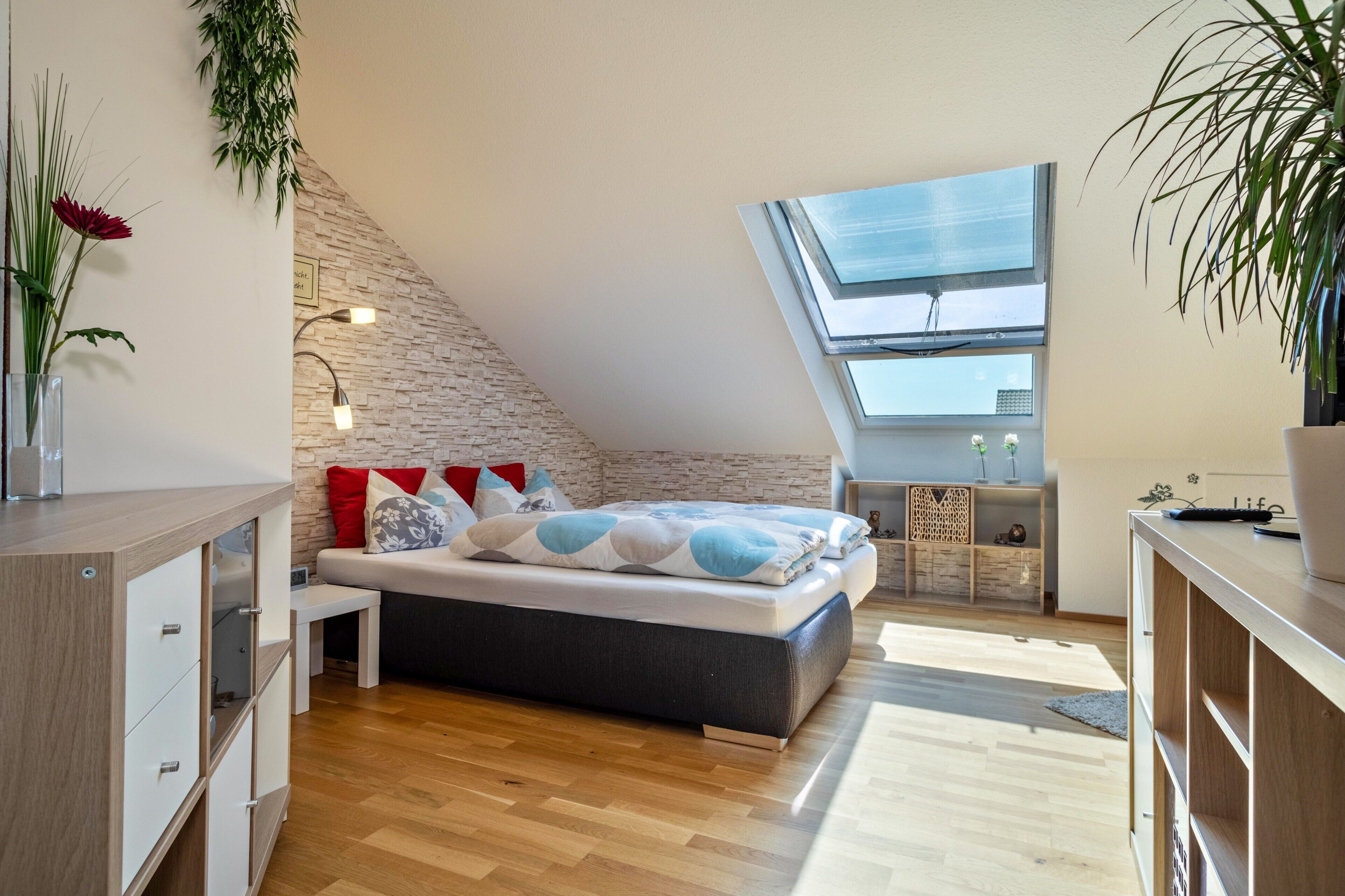 Une chambre moderne avec un plafond en pente, un puits de lumière, un lit avec une literie à motifs, une table de chevet et une commode en bois.
