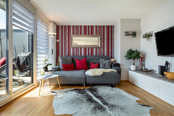 Un salon moderne avec un canapé gris, des coussins rouges, un grand miroir et un téléviseur à écran plat.